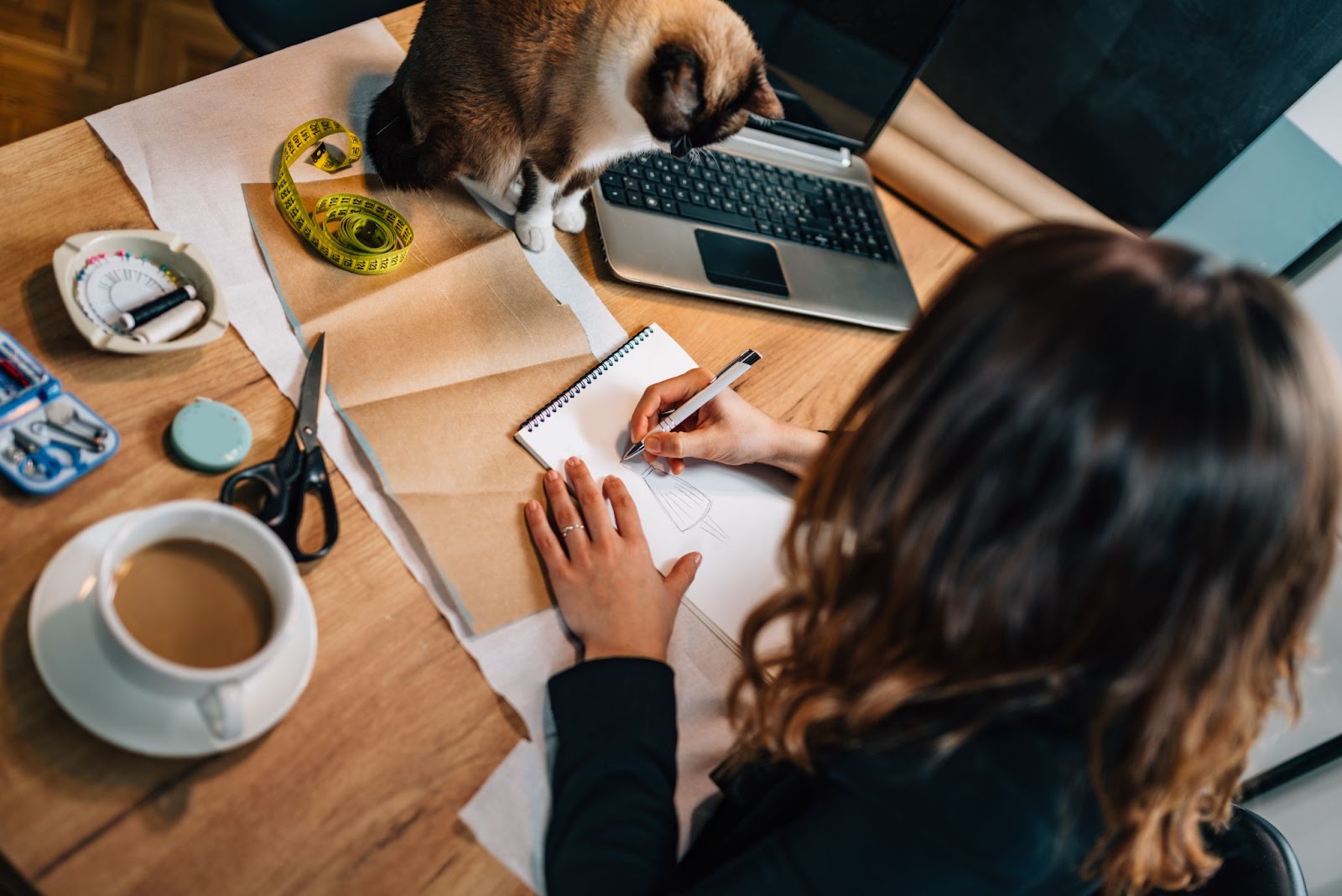 Mulher desenhando croqui de moda na mesa. Na mesa tem um molde de saia com uma fita métrica por cima, um gato sentando olhando, um computador, materiais de costura e uma xícara de café.